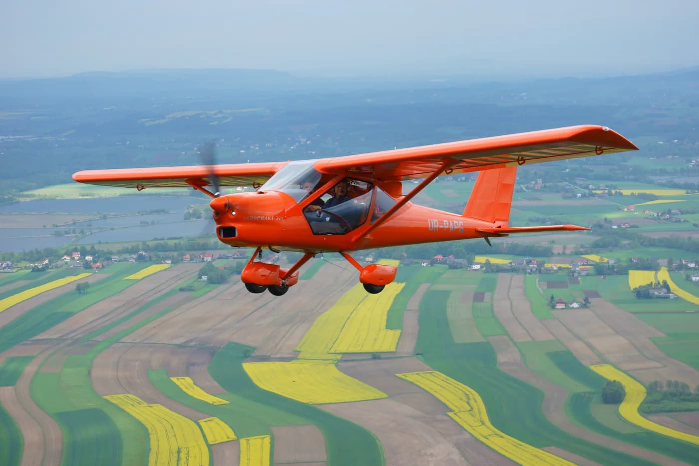 Red Aeroprakt A-32 photographed in flight over a patchwork of cultivated fields