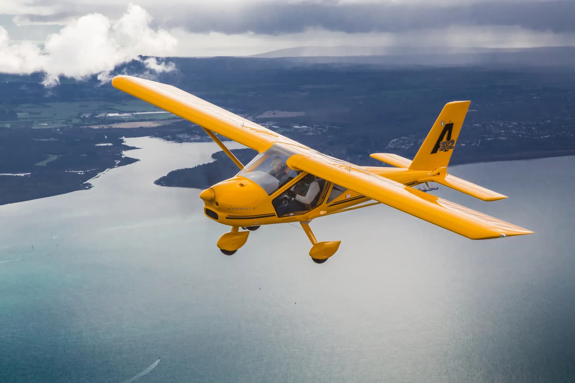 Yellow Aeroprakt A-32 flying above a winding waterway and wooded shoreline